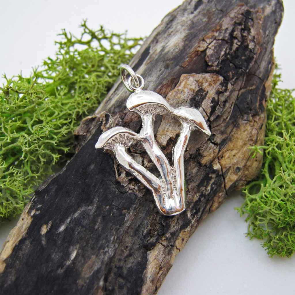A silver pendant of three mushrooms, resting on dark driftwood